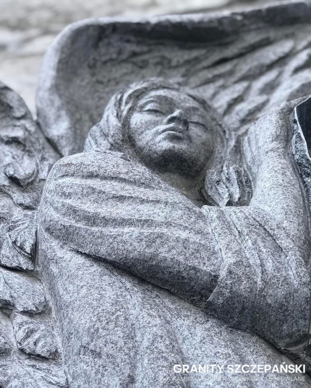 Granitowa rzeźba anioła z zamkniętymi oczami, detal nagrobka. Szary kamień z widoczną fakturą. Ujęcie z dołu podkreśla monumentalność.