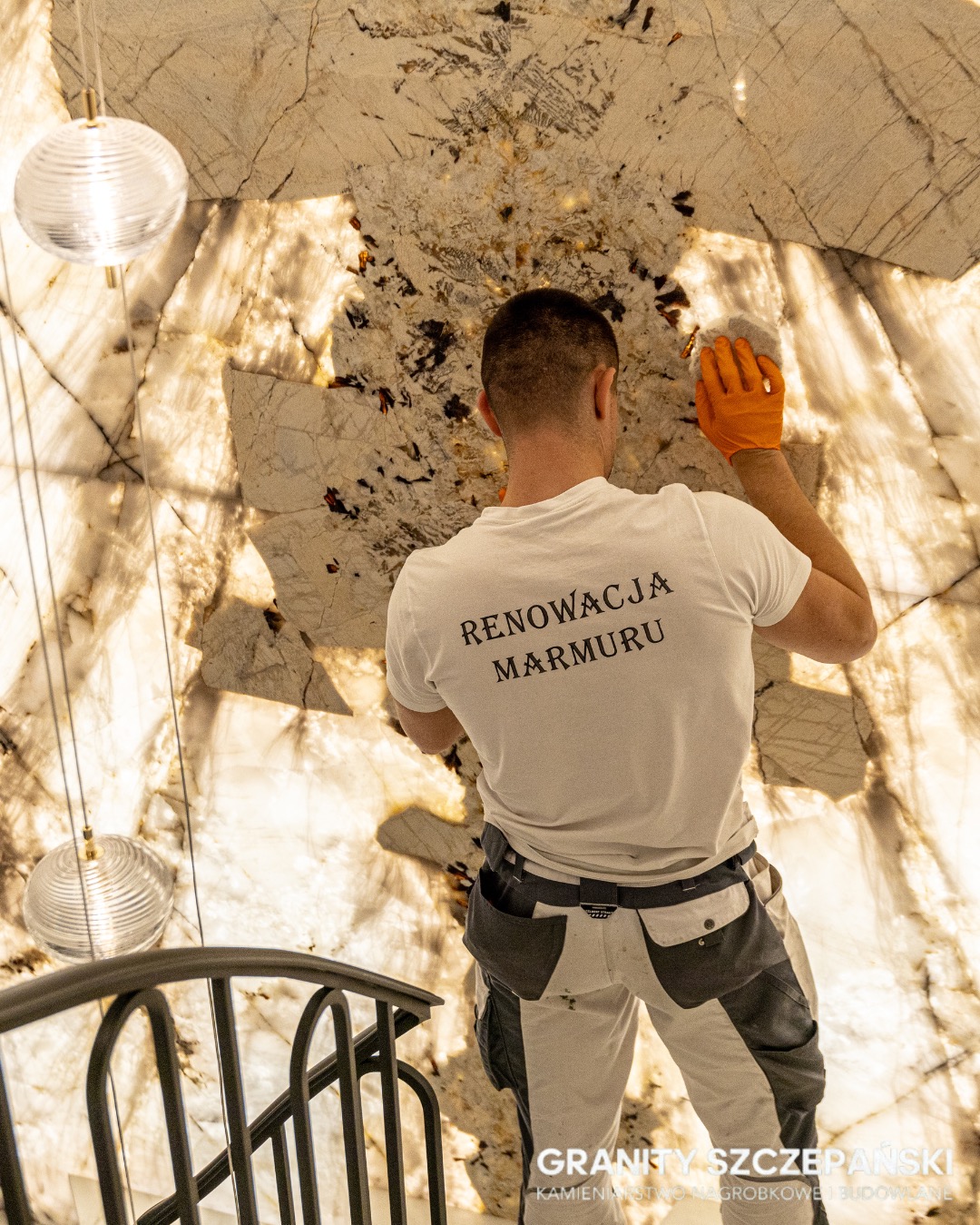 Pracownik w trakcie renowacji podświetlanej ściany z marmuru o unikalnym wzorze, widoczny od tyłu, z narzędziem w dłoni, oświetlenie dekoracyjne i fragment schodów.