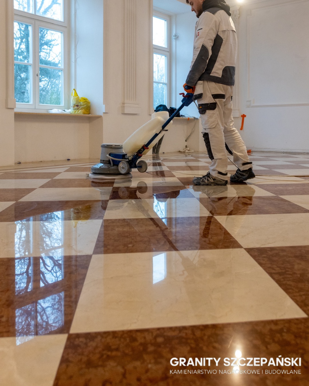 Polerowanie szachownicy z kamienia. Pracownik używa maszyny polerskiej, odbicia okien w wypolerowanej podłodze. Granity Szczepański.
