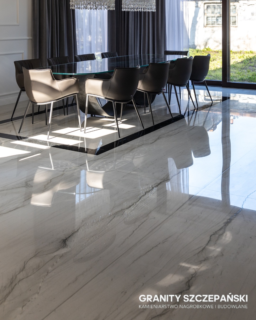 Polerowana podłoga z białego marmuru z szarymi żyłami, odbijająca światło i meble. W tle stół z krzesłami i okno z widokiem na zieleń.