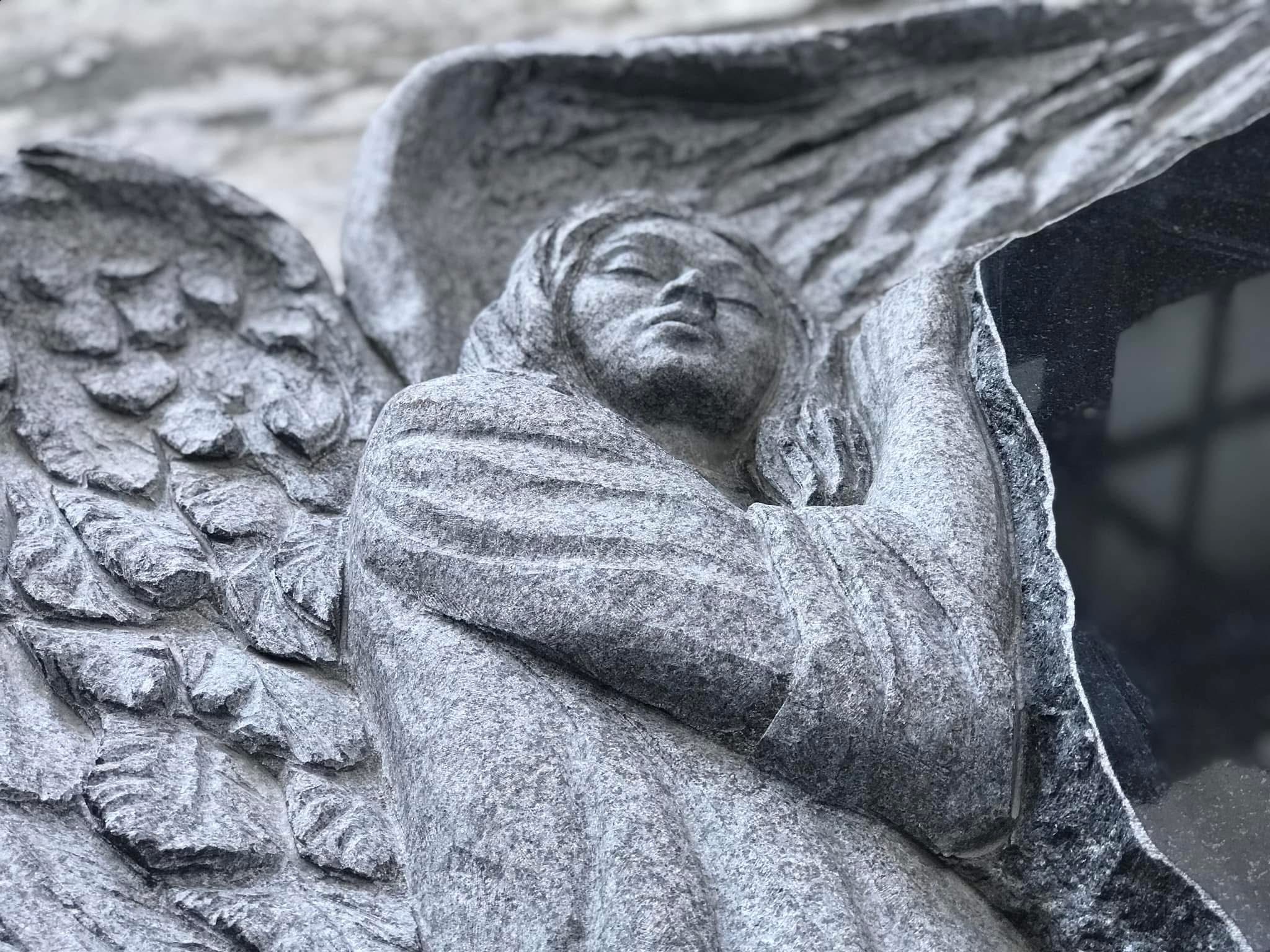 Granitowa rzeźba anioła z zamkniętymi oczami, część nagrobka z ciemnego kamienia. Delikatne światło podkreśla detale skrzydeł i draperii.