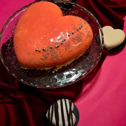 Adam Zygma - Pomarańczowe ciasto w kształcie serca na szklanym talerzu, ozdobione srebrnymi kuleczkami, obok ciastka i róży na tle czerwonej tkaniny i różowego tła.