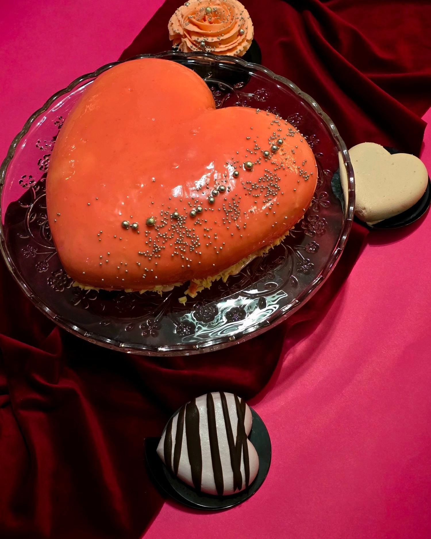 Pomarańczowe ciasto w kształcie serca na szklanym talerzu, ozdobione srebrnymi kuleczkami, obok ciastka i róży na tle czerwonej tkaniny i różowego tła.