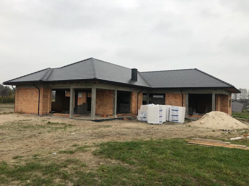 Dom w trakcie budowy z ciemnoszarym dachem z dachówki ceramicznej. Widoczne bloczki z betonu komórkowego i materiały budowlane. Pochmurne niebo w tle.