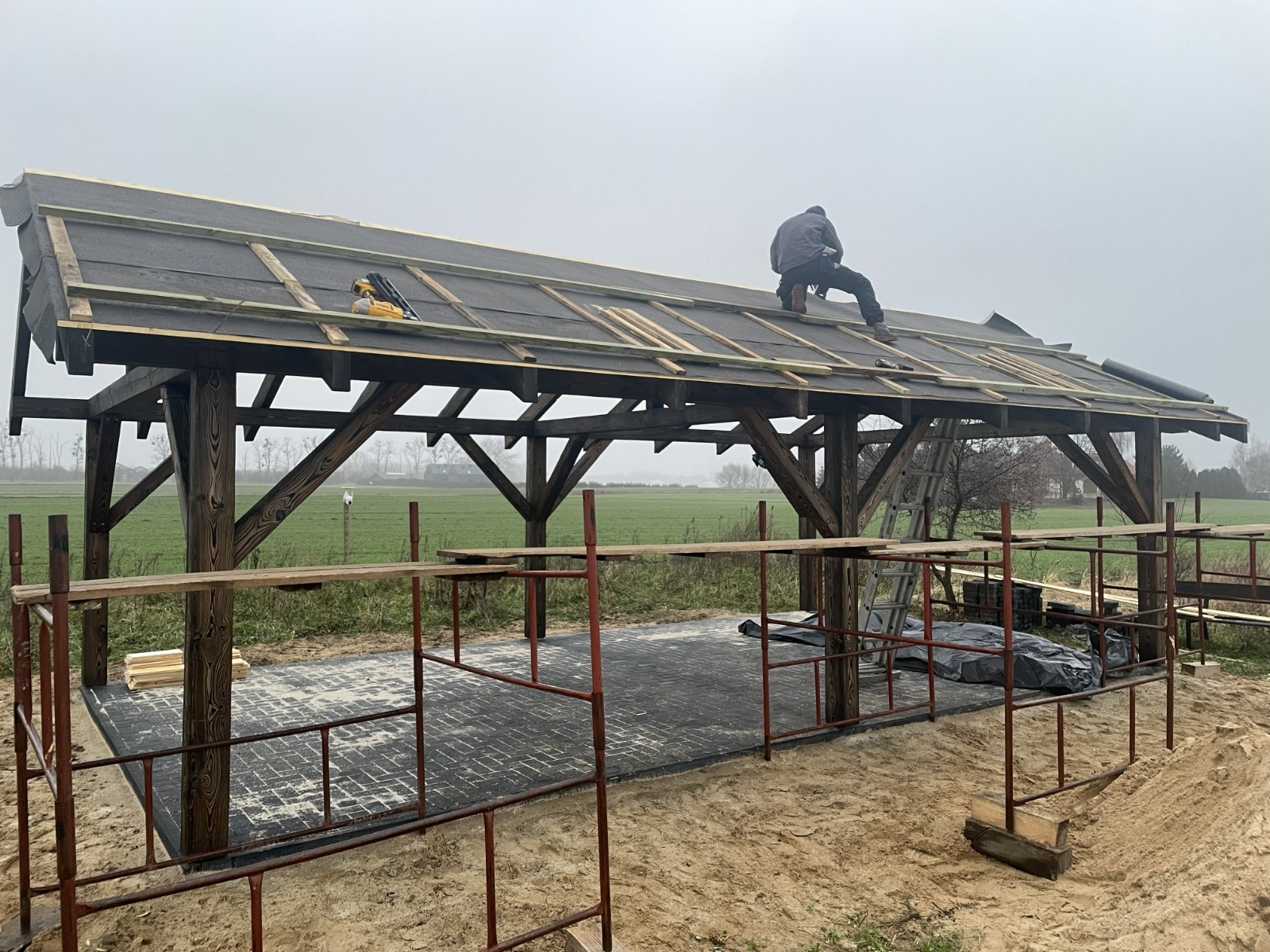 Montaż dachu na drewnianej konstrukcji z użyciem narzędzi. Widoczny pracownik na dachu i rusztowanie. Prace dekarskie w toku, z elementami izolacji i łat.