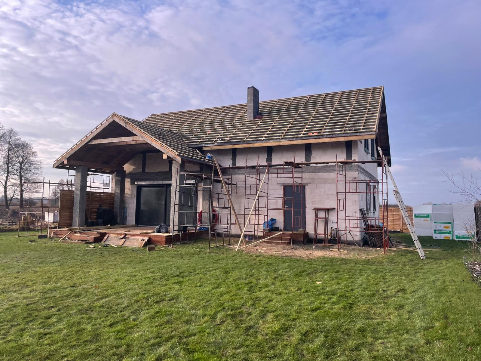 Budowa domu jednorodzinnego w trakcie realizacji. Widoczne rusztowania, ściany z bloczków i elementy konstrukcji dachu. Na pierwszym planie zielony trawnik.