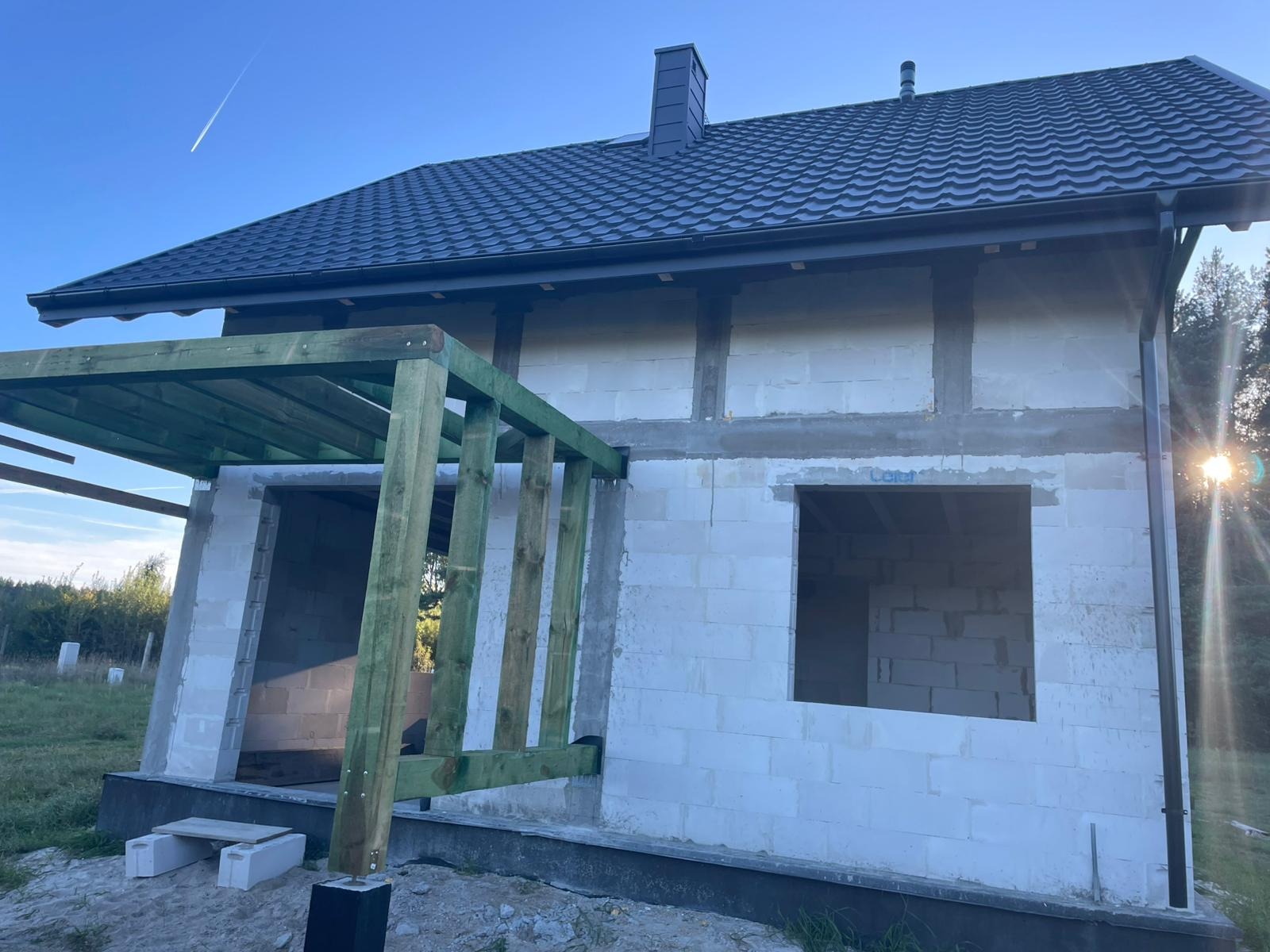 Dom w trakcie budowy z widoczną konstrukcją dachu z blachy trapezowej i drewnianą konstrukcją pergoli. Świecące słońce w tle.