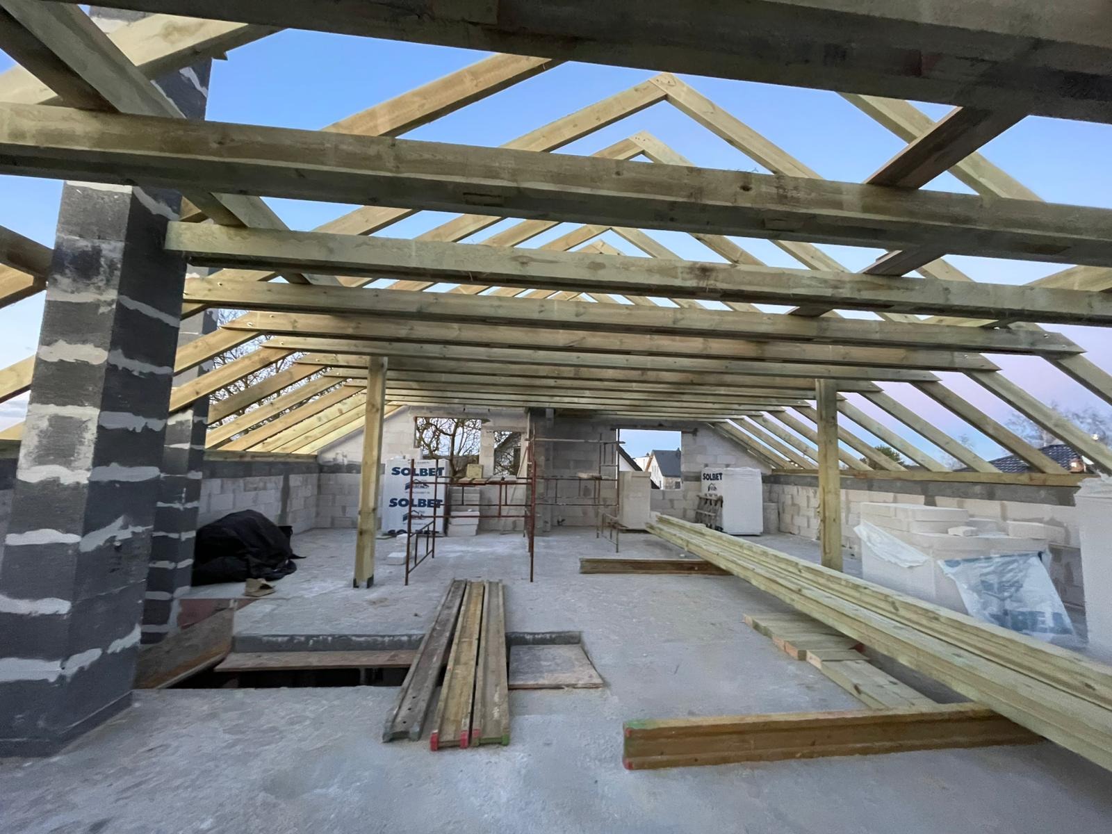 Wnętrze budynku w trakcie budowy z widoczną drewnianą konstrukcją dachu i elementami muru. Widoczne deski i materiały budowlane.