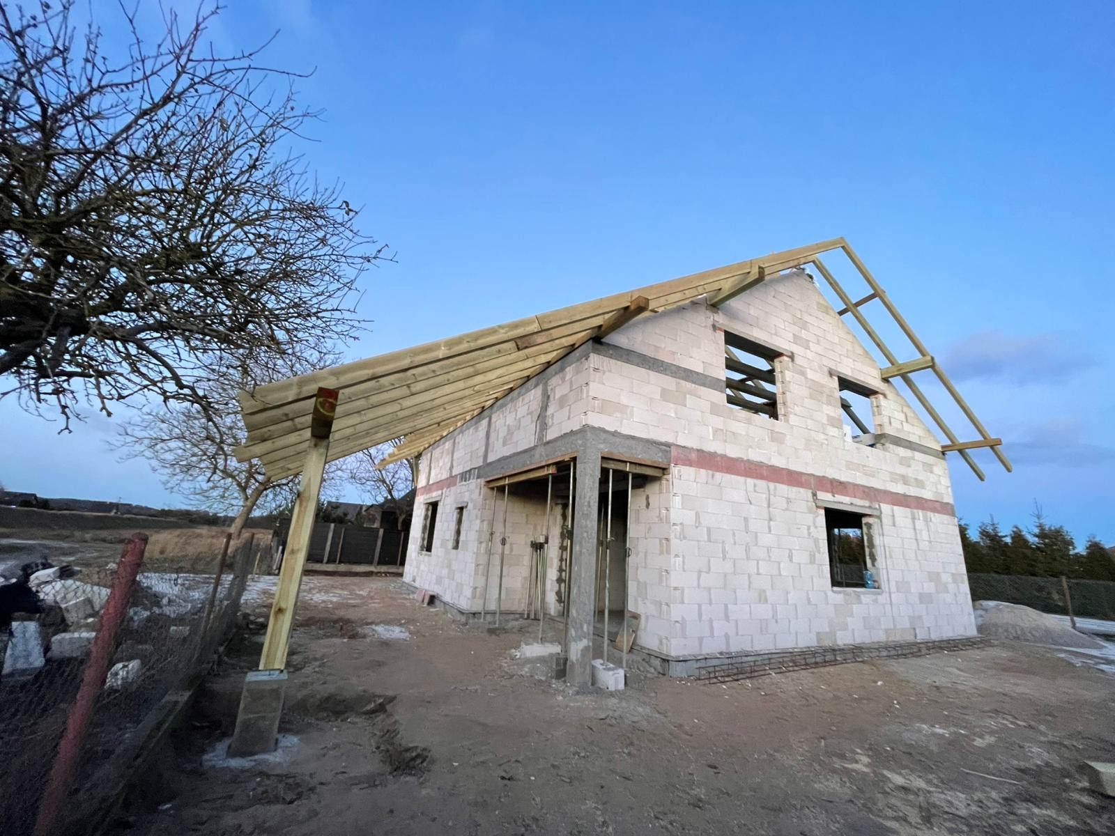Budowa domu z białych bloczków, widoczna konstrukcja dachu i zadaszenie tarasu. Surowy stan, w tle drzewo bez liści i błękitne niebo.