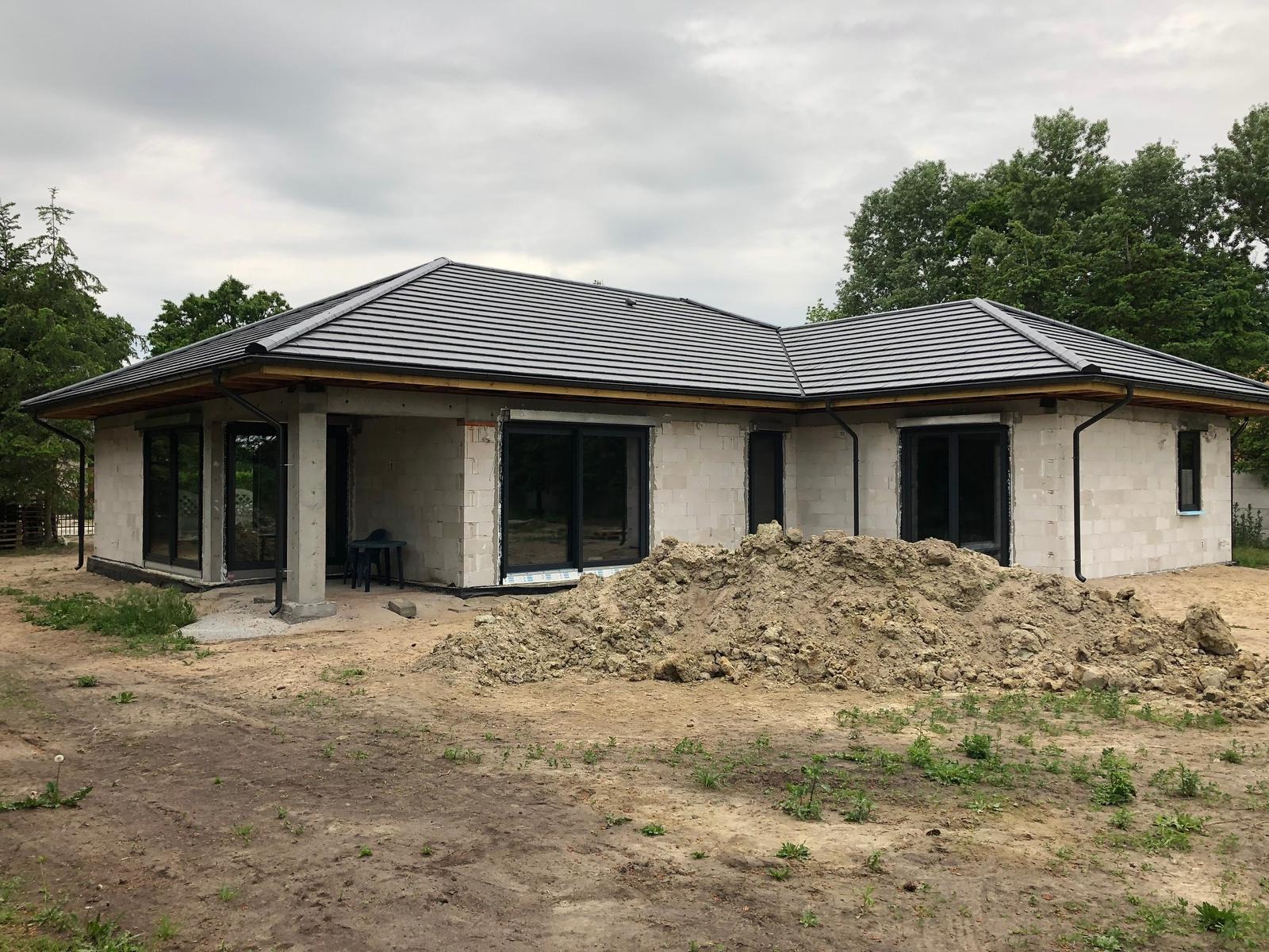 Dom jednorodzinny w trakcie budowy z ciemną dachówką. Widoczne okna z czarnymi ramami i sterta ziemi przed budynkiem. Pochmurne niebo w tle.