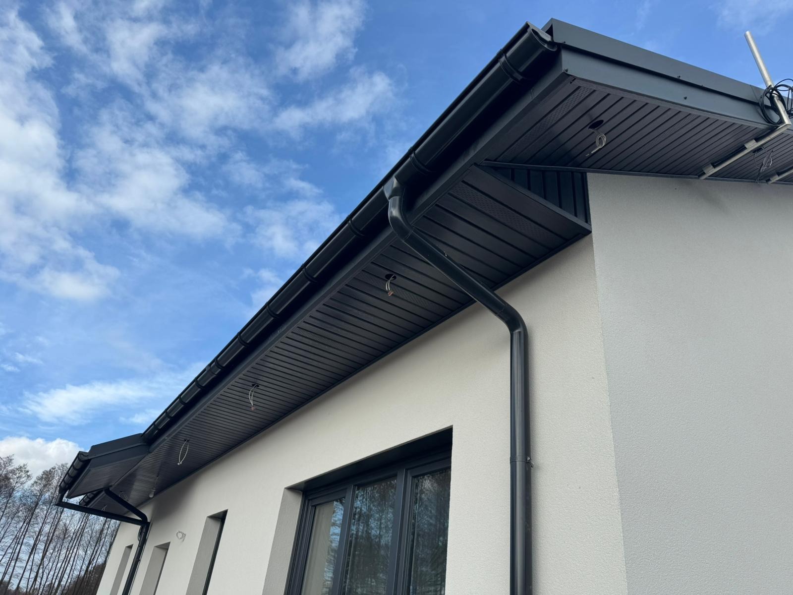Nowoczesny dom z grafitowym orynnowaniem i podbitką, widok z dołu na tle błękitnego nieba z chmurami. Detal architektoniczny, estetyczne wykończenie.