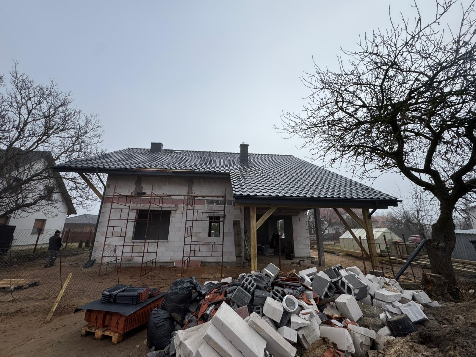 Dom w trakcie budowy z częściowo ukończonym dachem z ciemnej dachówki. Widoczne rusztowania, materiały budowlane i drewniana konstrukcja zadaszenia.