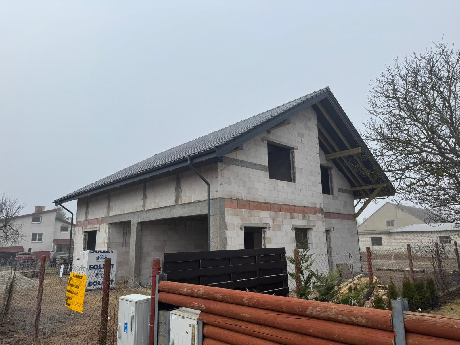 Dom w stanie surowym zamkniętym z szarą dachówką w Zławieś Mała. Widoczna więźba dachowa i okna bez szyb. Budynek z białego bloczka.