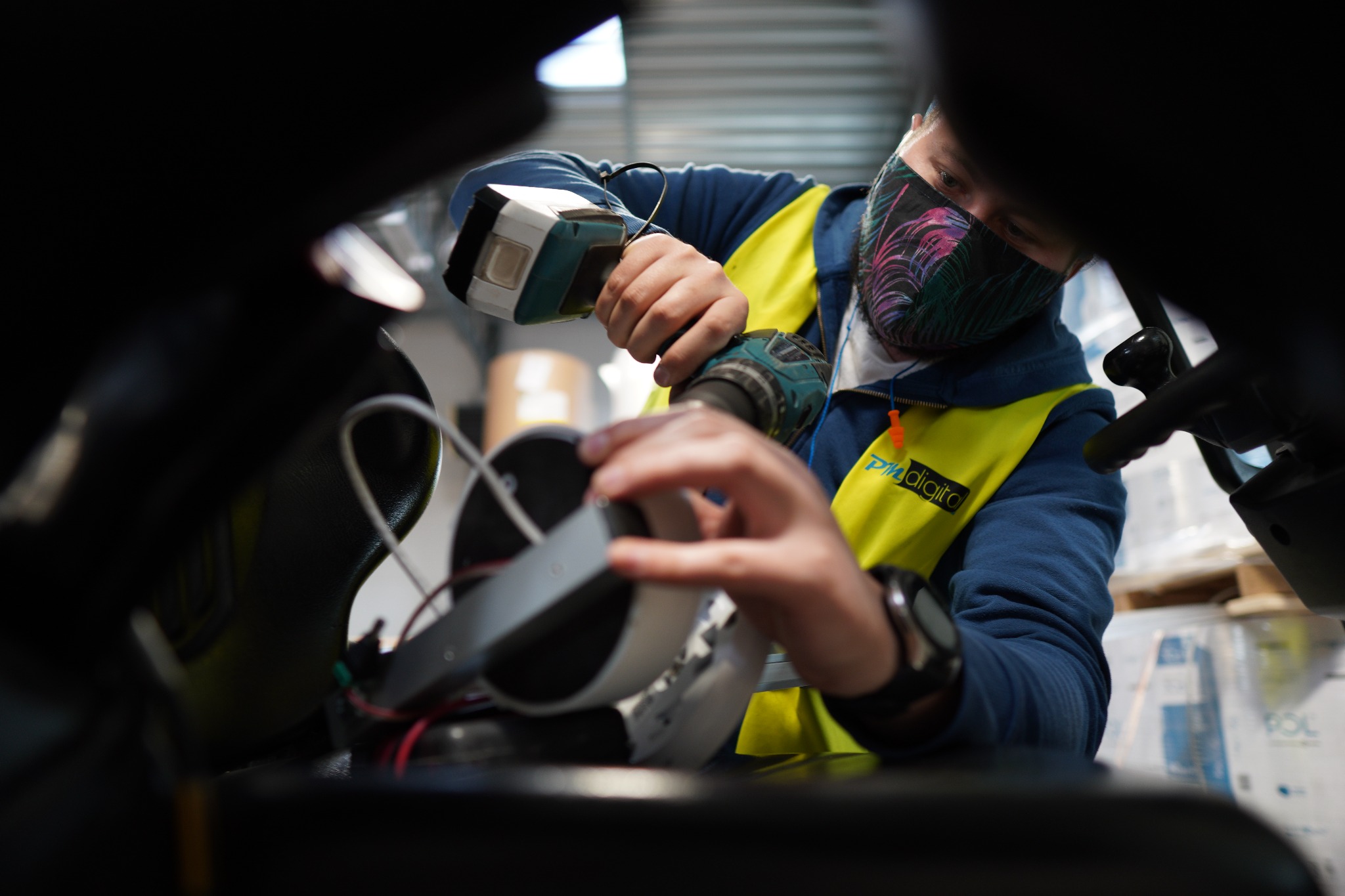 Instalator w masce, w kamizelce, montuje urządzenie z użyciem wkrętarki akumulatorowej. Widoczne przewody i elementy konstrukcyjne.