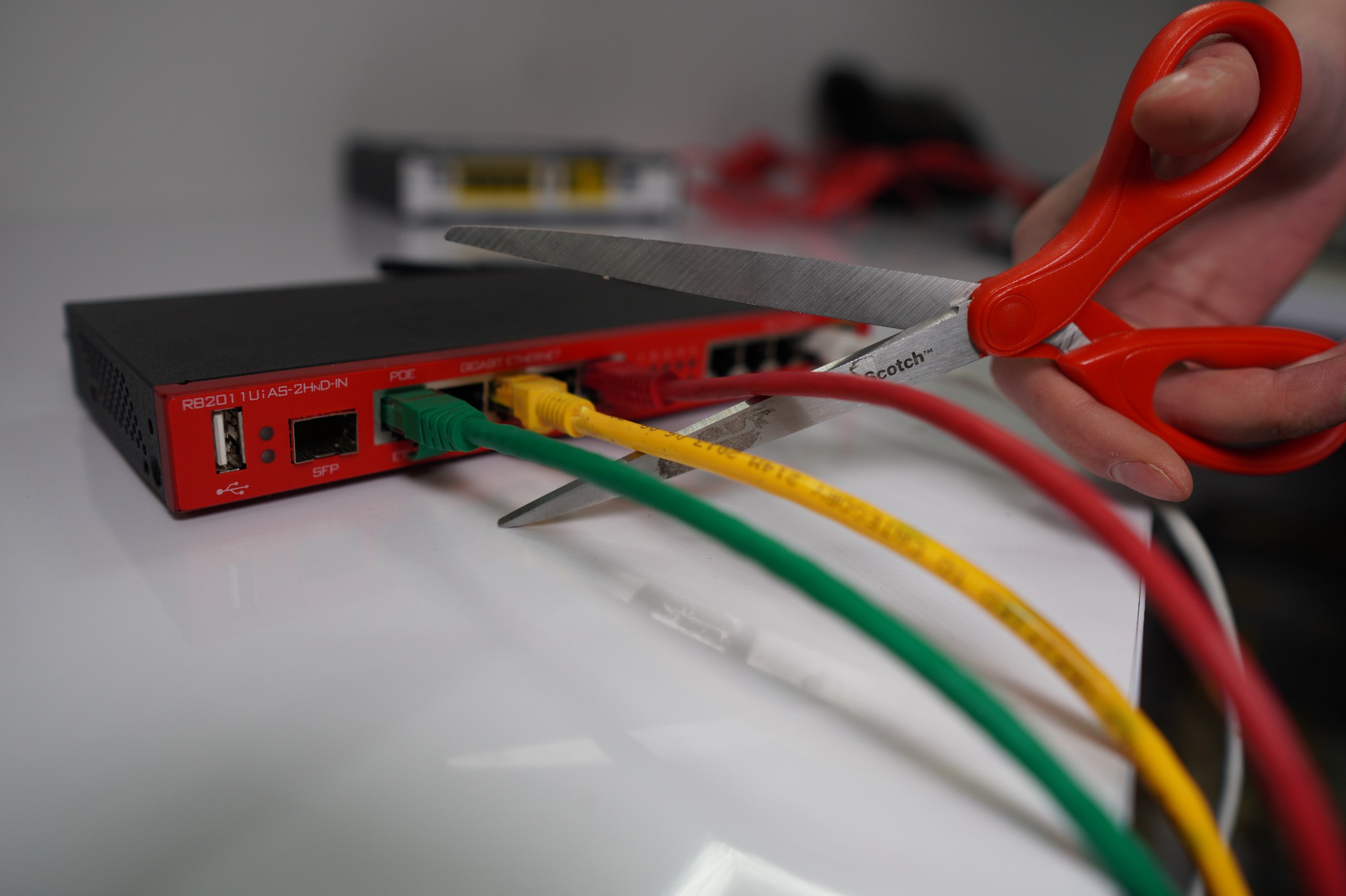 Czerwony router RB2011UiAS-2HnD-IN z podłączonymi kablami sieciowymi (zielony, żółty, czerwony) przecinanymi czerwonymi nożyczkami Scotch.