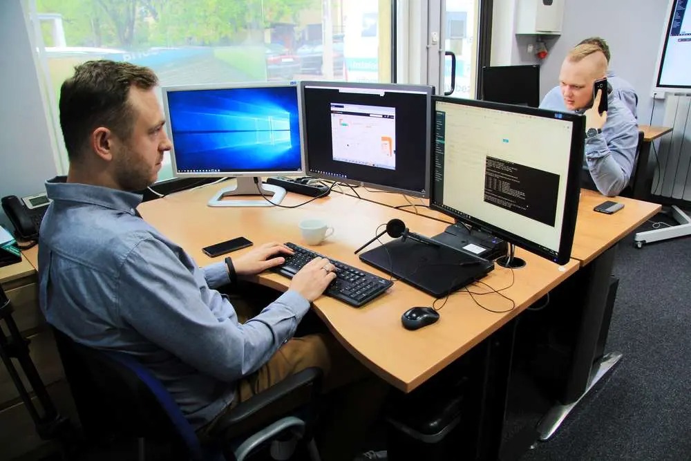 Pracownik IT przy biurku z trzema monitorami, klawiaturą i słuchawkami. Drugi pracownik rozmawia przez telefon w tle. Jasne biuro z oknem.