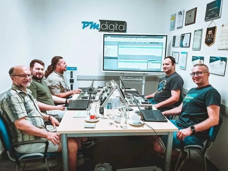 Zespół PM Digital pracuje przy biurkach z laptopami, analizując dane na dużym ekranie. Na ścianie wiszą certyfikaty i nagrody firmy.