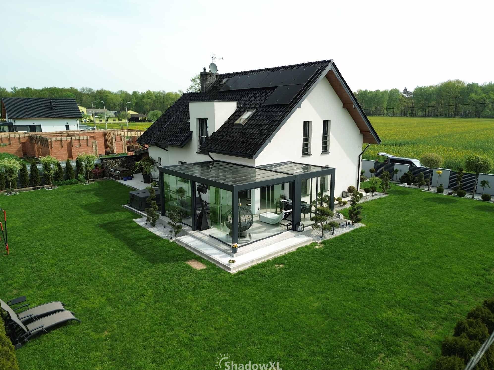 Szklana zabudowa balkonu z czarną ramą, tworząca nowoczesną werandę QubeXL w Żorach. Widok z góry na narożną konstrukcję i zadbany trawnik.