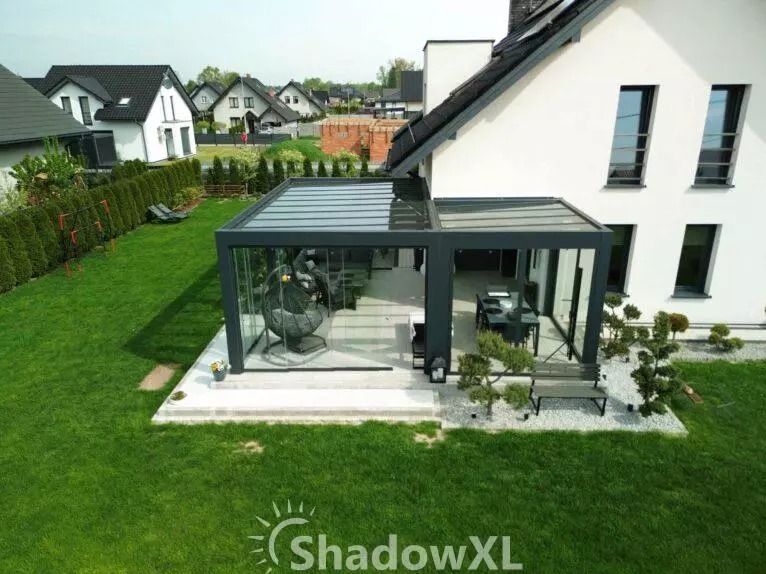 Nowoczesna, przeszklona zabudowa balkonu z metalową konstrukcją, widok z góry na tle zadbanego ogrodu i domów jednorodzinnych. Wewnątrz meble ogrodowe.