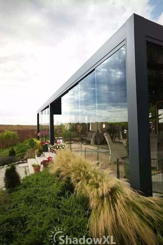 Szklana zabudowa tarasu z metalową ramą, widok z zewnątrz na ogród. W tle meble ogrodowe i roślinność. Nowoczesny design.