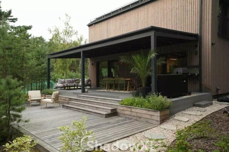 Nowoczesna, czarna pergola z płaskim dachem, przylegająca do drewnianego domu. Taras z meblami ogrodowymi, rośliny w donicach. Widok na ogród.