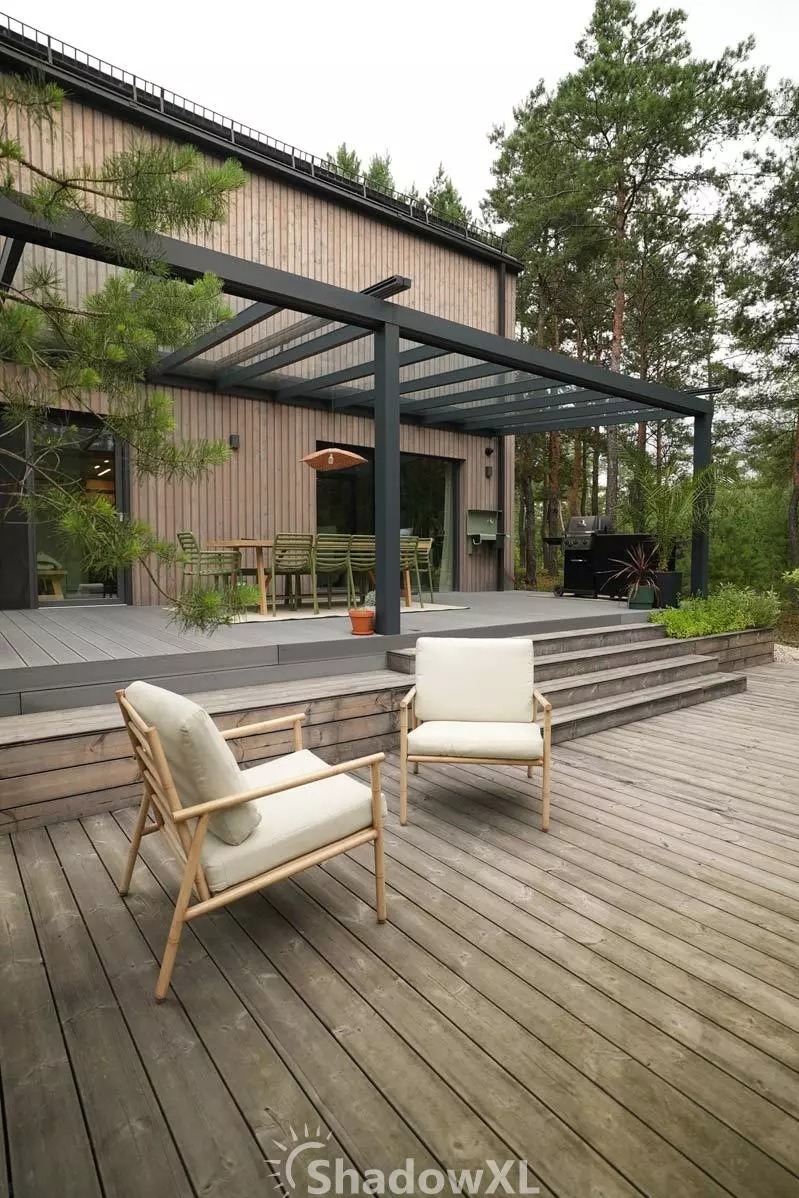 Nowoczesna, grafitowa pergola z przezroczystym zadaszeniem, przylegająca do drewnianego domu. Dwa kremowe fotele na tarasie, zieleń w tle.