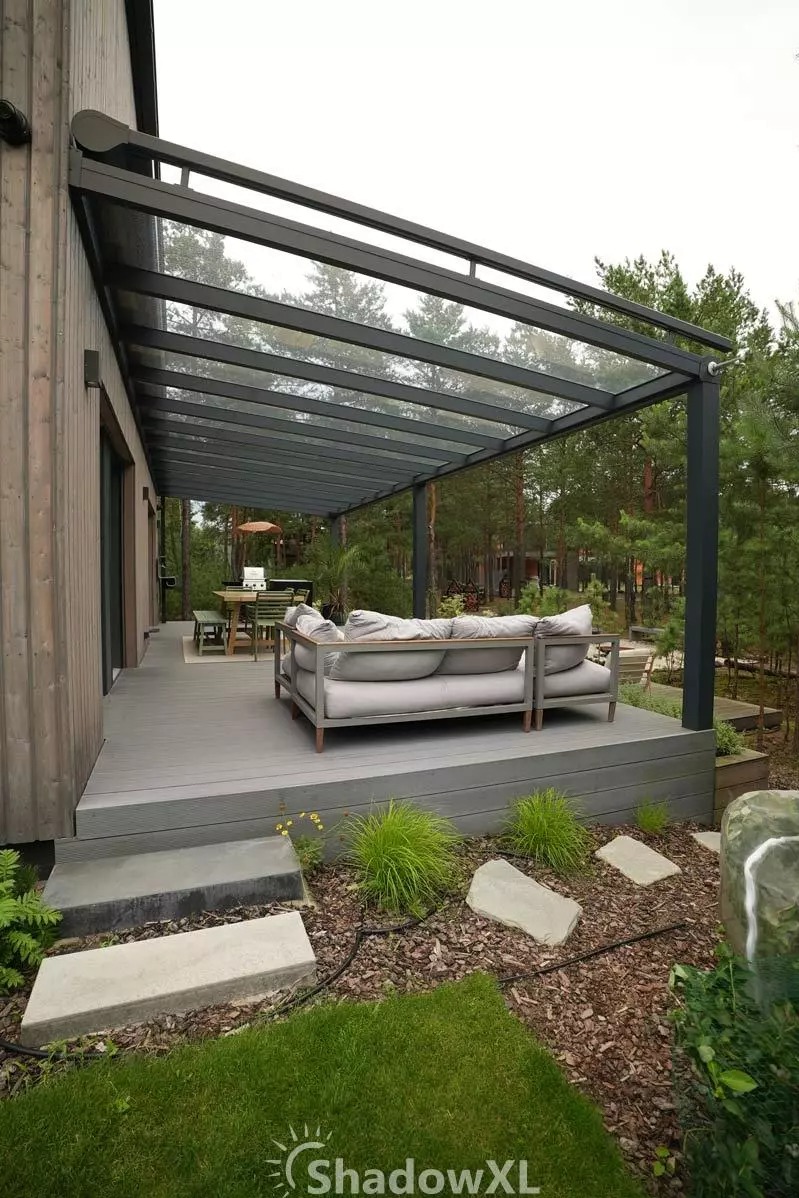Szklana pergola z metalową konstrukcją nad tarasem z szarą sofą i stołem jadalnym. Widok na ogród z drzewami w tle. Nowoczesny design.
