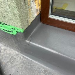 PRO DACHY - Hydroizolacje - Szczegółowe ujęcie hydroizolacji okna dachowego z użyciem taśmy uszczelniającej, widoczne połączenie z ociepleniem ściany. Szara powierzchnia hydroizolacji.