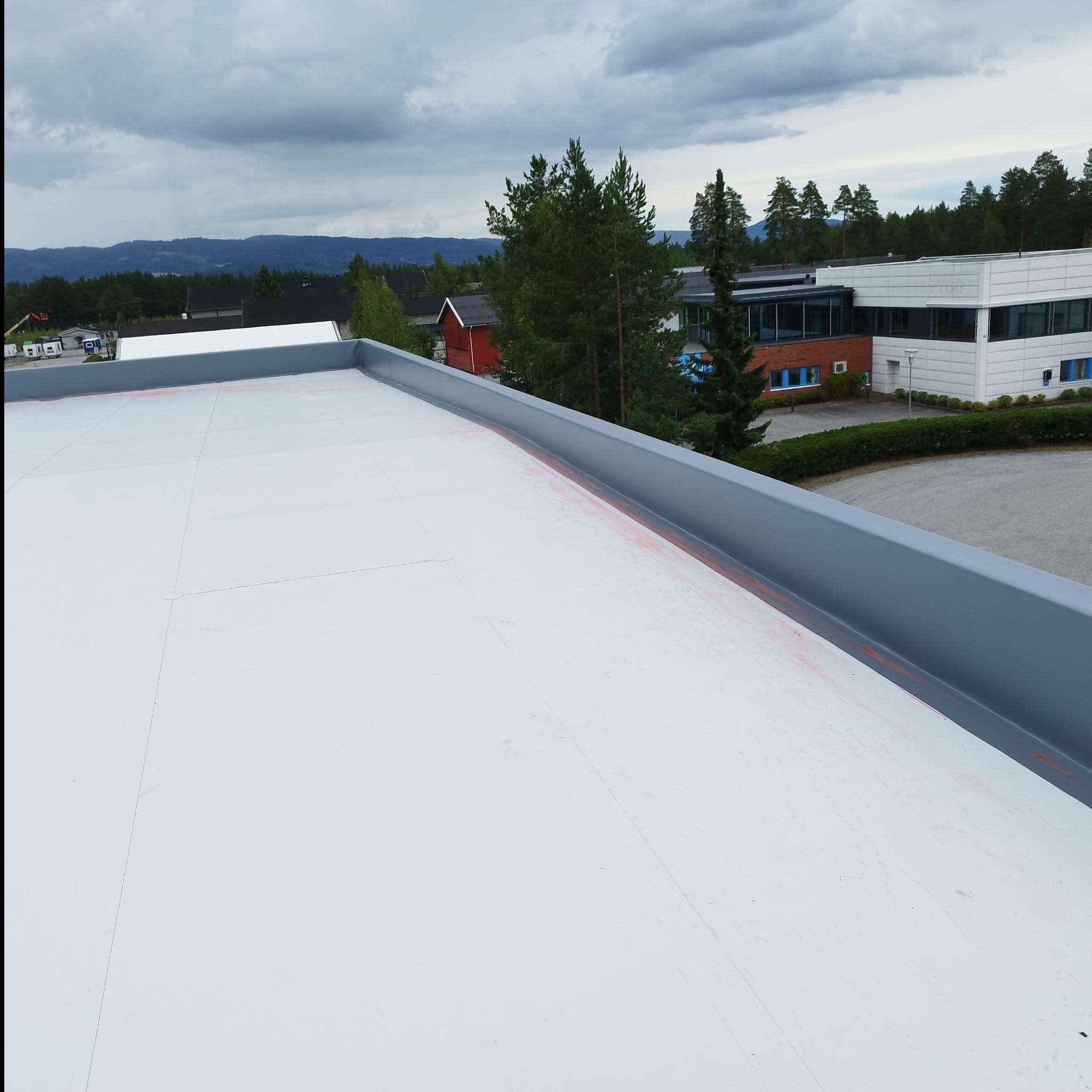 Biała, płaska powierzchnia dachu z szarą obróbką blacharską na krawędzi, w tle budynki i drzewa pod pochmurnym niebem. Widok z góry.