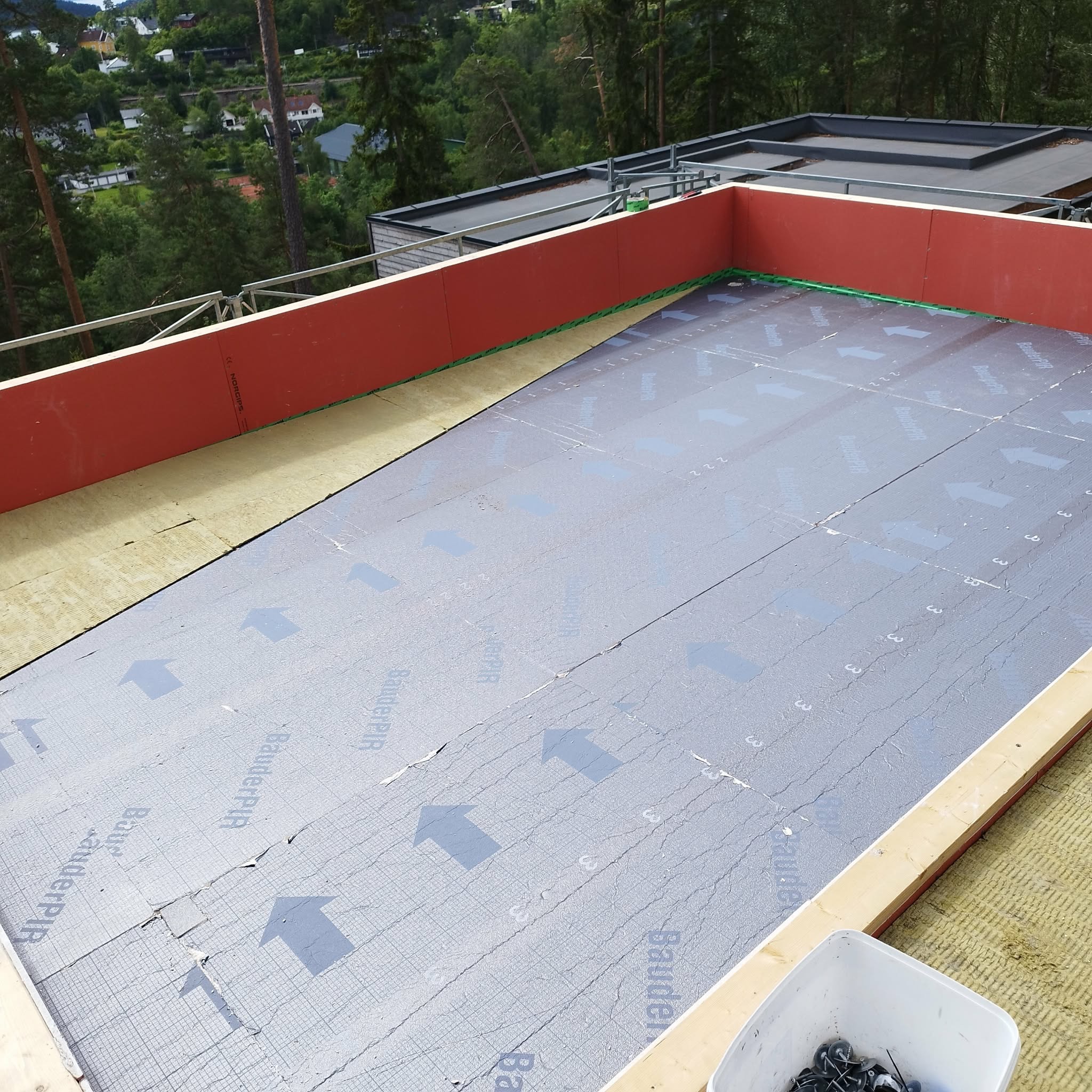 Dach w trakcie krycia: widoczna izolacja, membrana z nadrukiem strzałek i częściowo zamontowane czerwone panele. W tle zieleń drzew i zabudowania.