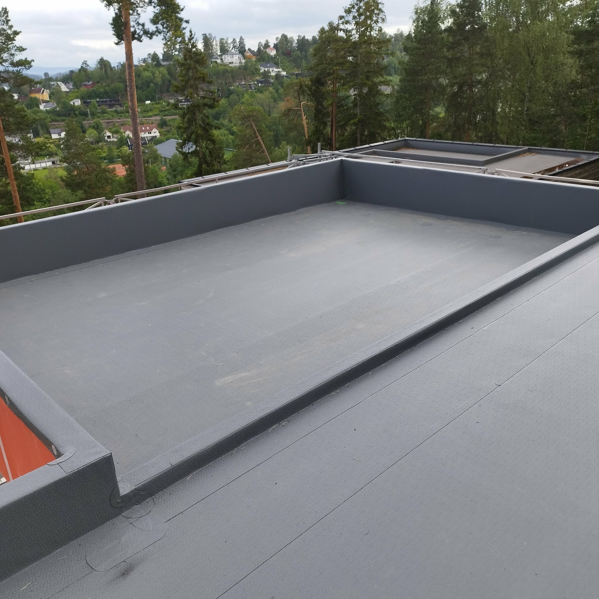 Szary, płaski dach z obróbkami blacharskimi, widok z góry. W tle zieleń drzew i zabudowania mieszkalne. Nowoczesne wykończenie dachu.