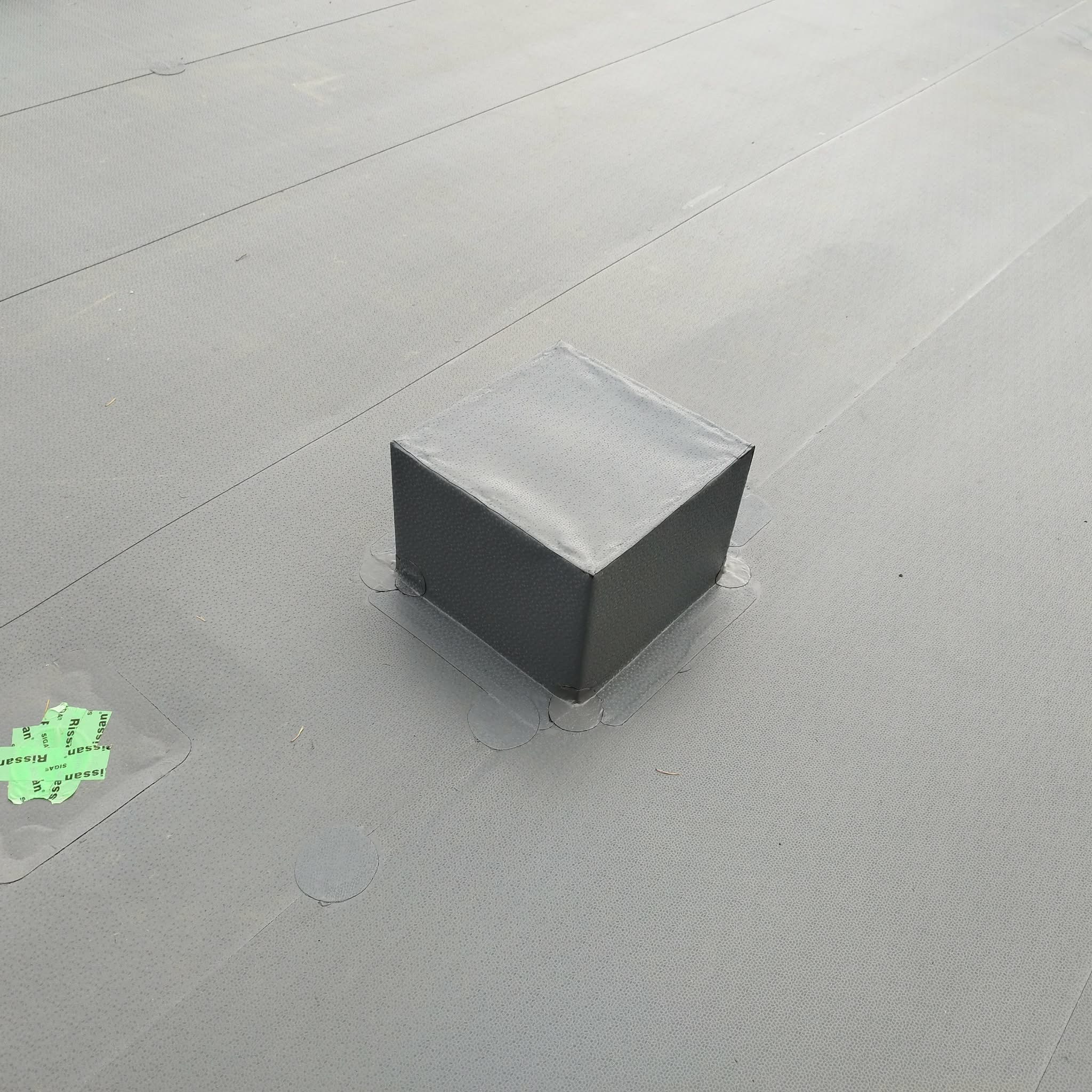 Szary, płaski dach z pojedynczym, kwadratowym kominkiem wentylacyjnym. Widoczne łączenia papy i naklejka z logo producenta. Ujęcie z góry.