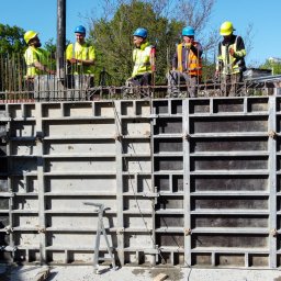 PRATA Sp. z o. o. - Ekipa budowlana w kaskach nad szalunkiem zbrojeniowym, przygotowująca się do wylewania betonu. Widoczny dźwig do betonu i zieleń w tle.