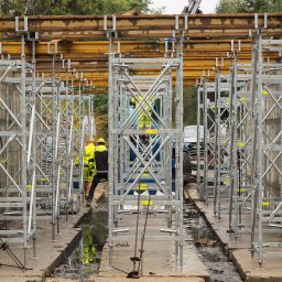 PRATA Sp. z o. o. - Szare, metalowe rusztowania budowlane z żółtymi elementami, ustawione w rzędzie na placu budowy. W tle widoczni pracownicy w odblaskowych ubraniach i pojazd.