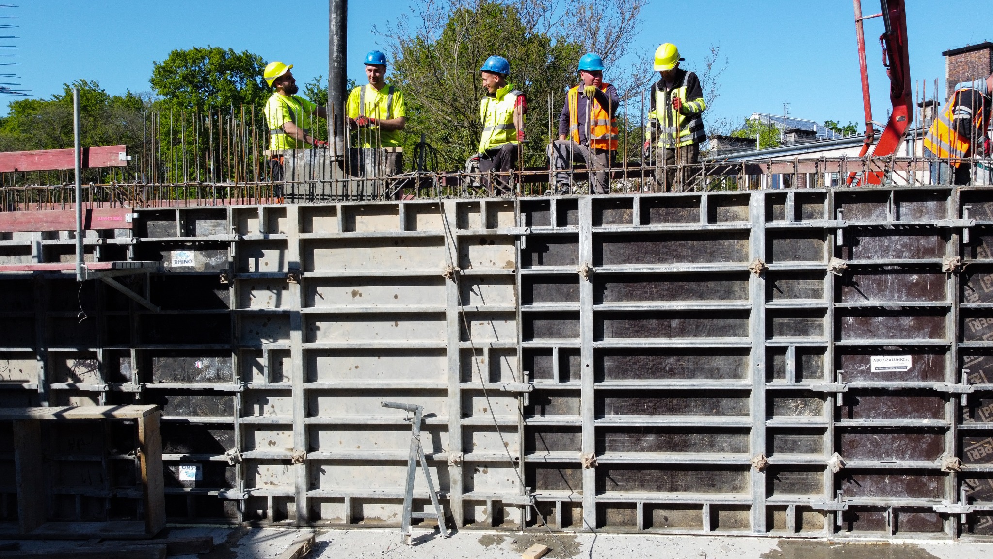 Ekipa budowlana w kaskach nad szalunkiem zbrojeniowym, przygotowująca się do wylewania betonu. Widoczny dźwig do betonu i zieleń w tle.