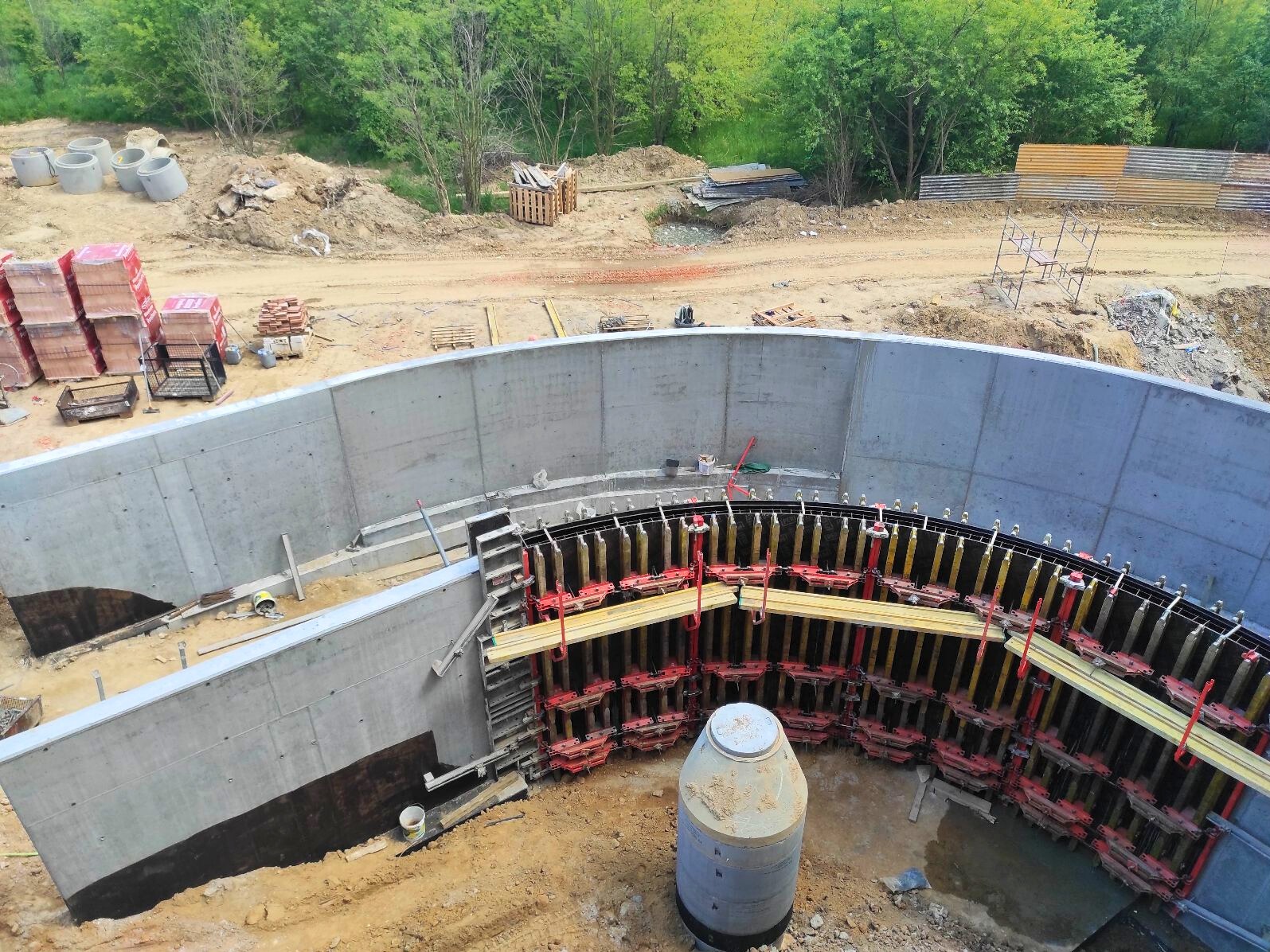 Budowa betonowego zbiornika retencyjnego: szalunki łukowe, betonowe ściany, rury betonowe. Widok z góry na plac budowy w otoczeniu zieleni.