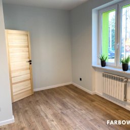 Farbowo - Jasne, pomalowane na szaro wnętrze pokoju z drewnianą podłogą i oknem z zieloną ramą. Widoczny grzejnik i drewniane drzwi. Minimalistyczny design.