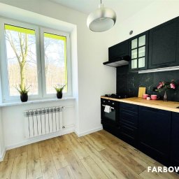 Farbowo - Nowoczesna kuchnia po odświeżeniu: czarne szafki kontrastują z drewnianym blatem i jasną podłogą. Ściany białe, okno z zielonym akcentem. Minimalistyczny design.