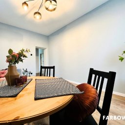 Farbowo - Jasne wnętrze po odświeżeniu: okrągły stół z dekoracjami, czarne krzesła i subtelne, szare ściany. Nowoczesna lampa sufitowa i roślina doniczkowa dopełniają aranżację.