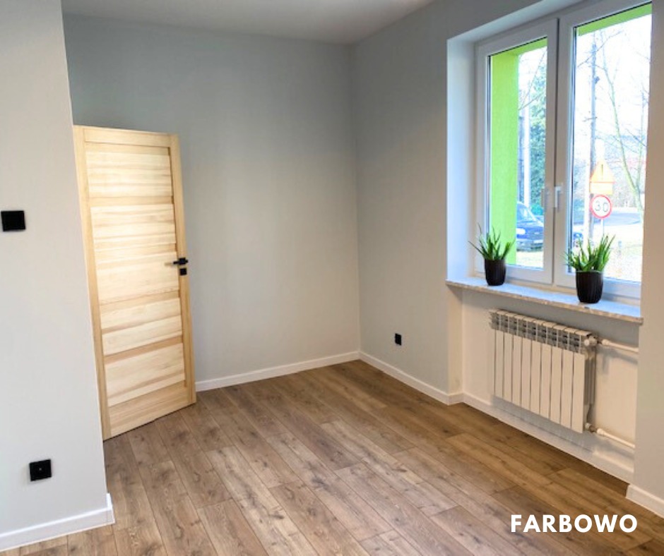 Jasne, pomalowane na szaro wnętrze pokoju z drewnianą podłogą i oknem z zieloną ramą. Widoczny grzejnik i drewniane drzwi. Minimalistyczny design.