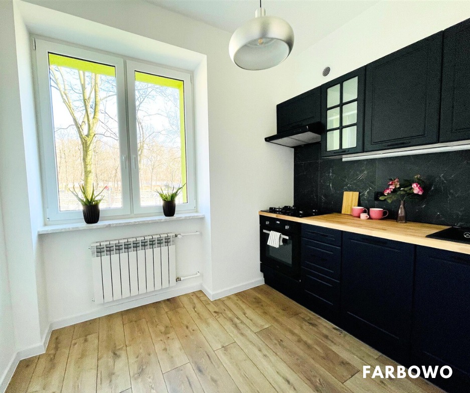 Nowoczesna kuchnia po odświeżeniu: czarne szafki kontrastują z drewnianym blatem i jasną podłogą. Ściany białe, okno z zielonym akcentem. Minimalistyczny design.