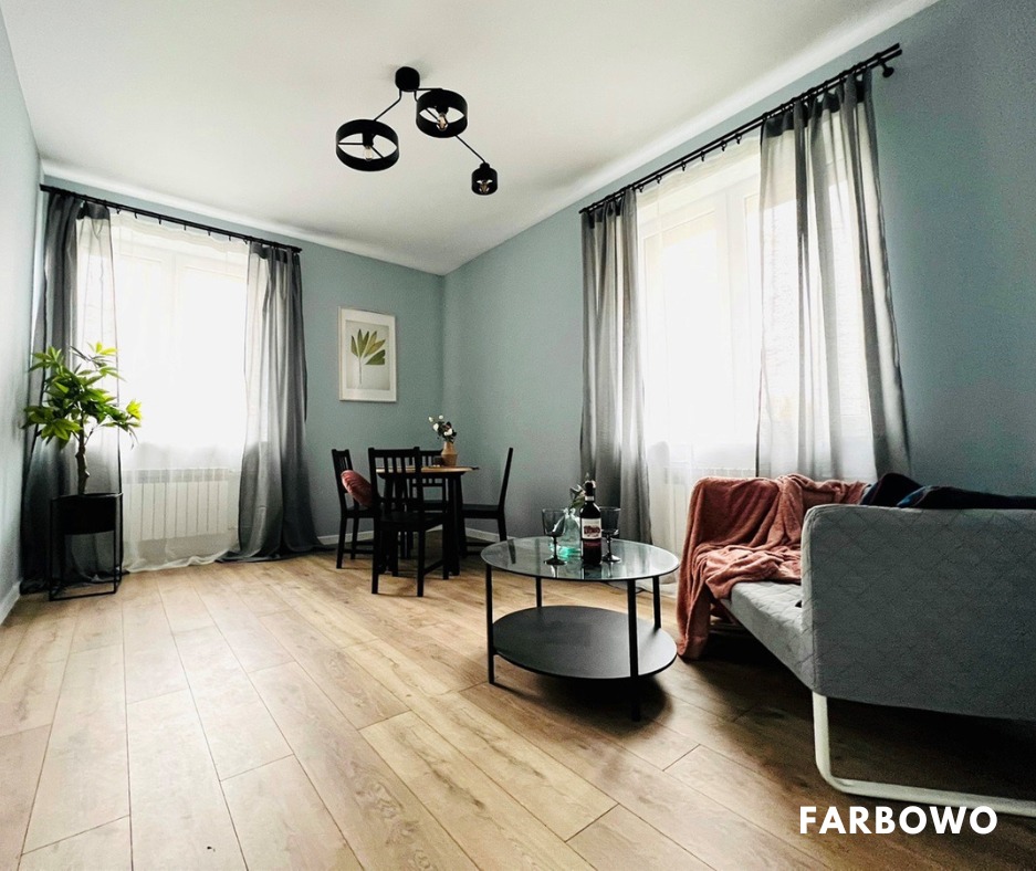 Jasne wnętrze po odświeżeniu: błękitne ściany, drewniana podłoga, czarne meble i lampa. Stonowana kolorystyka, minimalistyczny wystrój salonu z oknami.