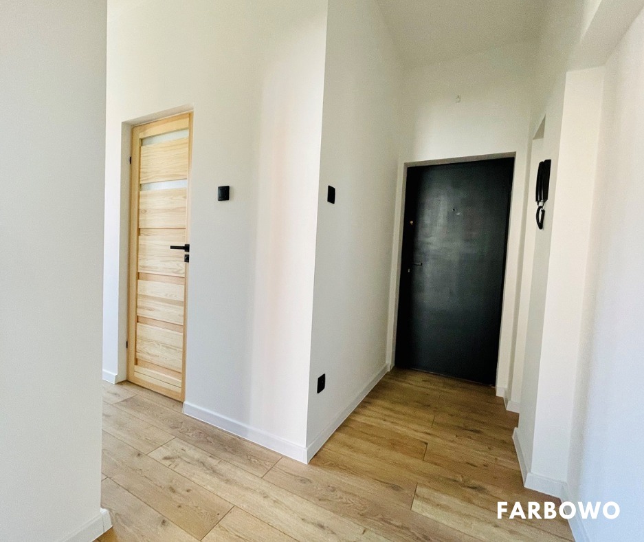 Jasne wnętrze po odświeżeniu: białe ściany, drewniana podłoga i drzwi, czarne akcenty w postaci kontaktów i drzwi wejściowych. Minimalistyczny design.