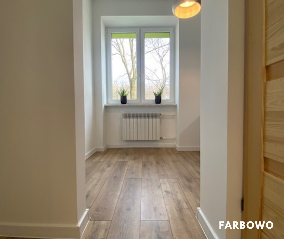 Jasne wnętrze po remoncie: białe ściany, drewniana podłoga, okno z widokiem na drzewa. Minimalistyczny design, czyste linie, świeżo pomalowane ściany.