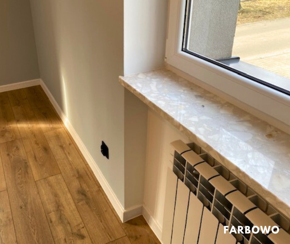 Jasny pokój po malowaniu: świeżo pomalowane ściany w odcieniach szarości, drewniana podłoga, marmurowy parapet i biały grzejnik. Widoczne gniazdko elektryczne.