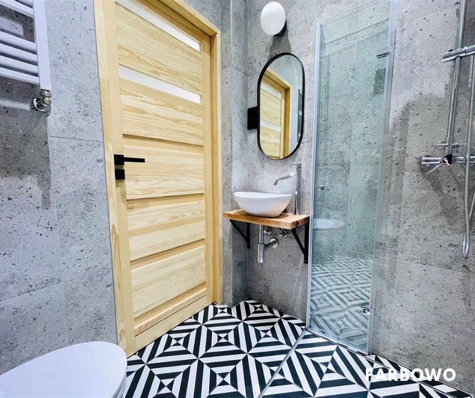 Wykończona łazienka z drewnianymi drzwiami, umywalką na drewnianym blacie i kabiną prysznicową. Szaro-biała kolorystyka, wzorzysta podłoga.