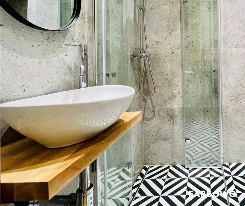 Nowoczesna łazienka z białą umywalką na drewnianym blacie, szklaną kabiną prysznicową i geometryczną podłogą w czarno-białe wzory. Ściany wykończone płytkami imitującymi beton.