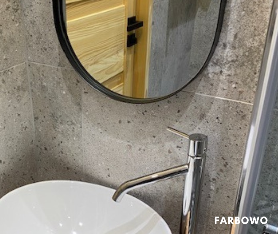 Wykończona łazienka z umywalką, baterią i lustrem w czarnej ramie. Widoczne szare płytki na ścianie i fragment kabiny prysznicowej. W lustrze odbija się drewniane wykończenie.