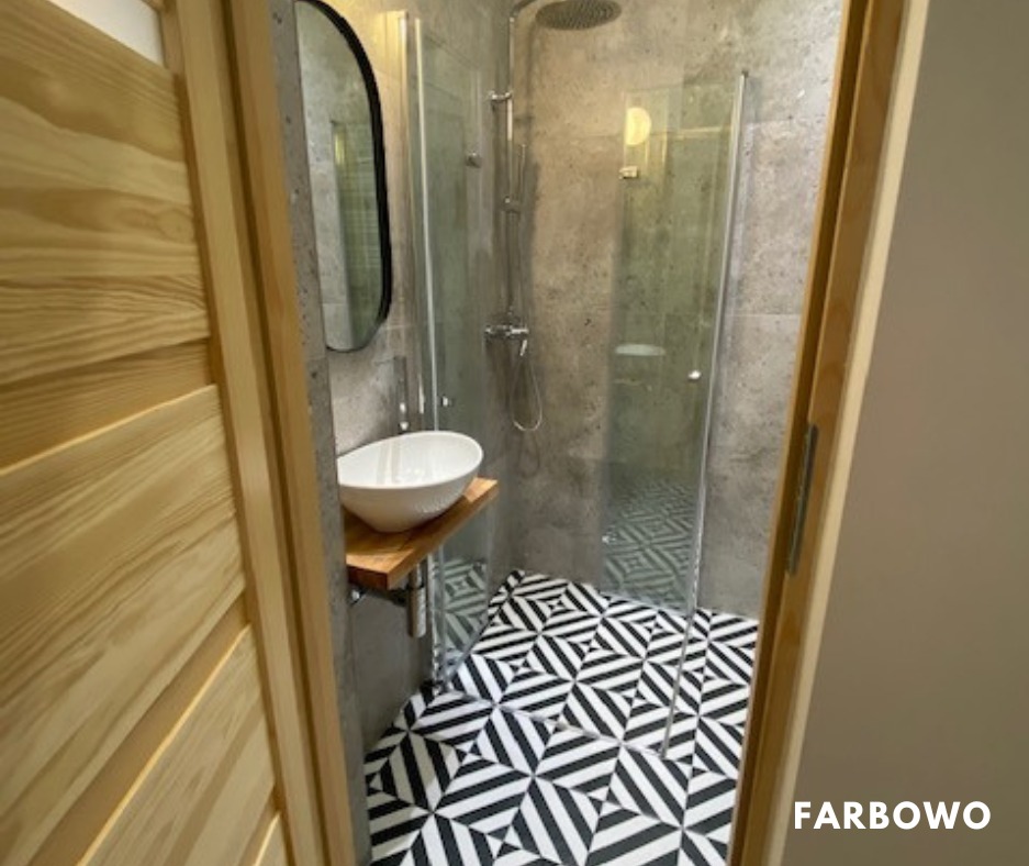 Nowoczesna łazienka z kabiną prysznicową, umywalką na drewnianym blacie i czarno-białą podłogą w geometryczny wzór, widziana zza drewnianych drzwi.