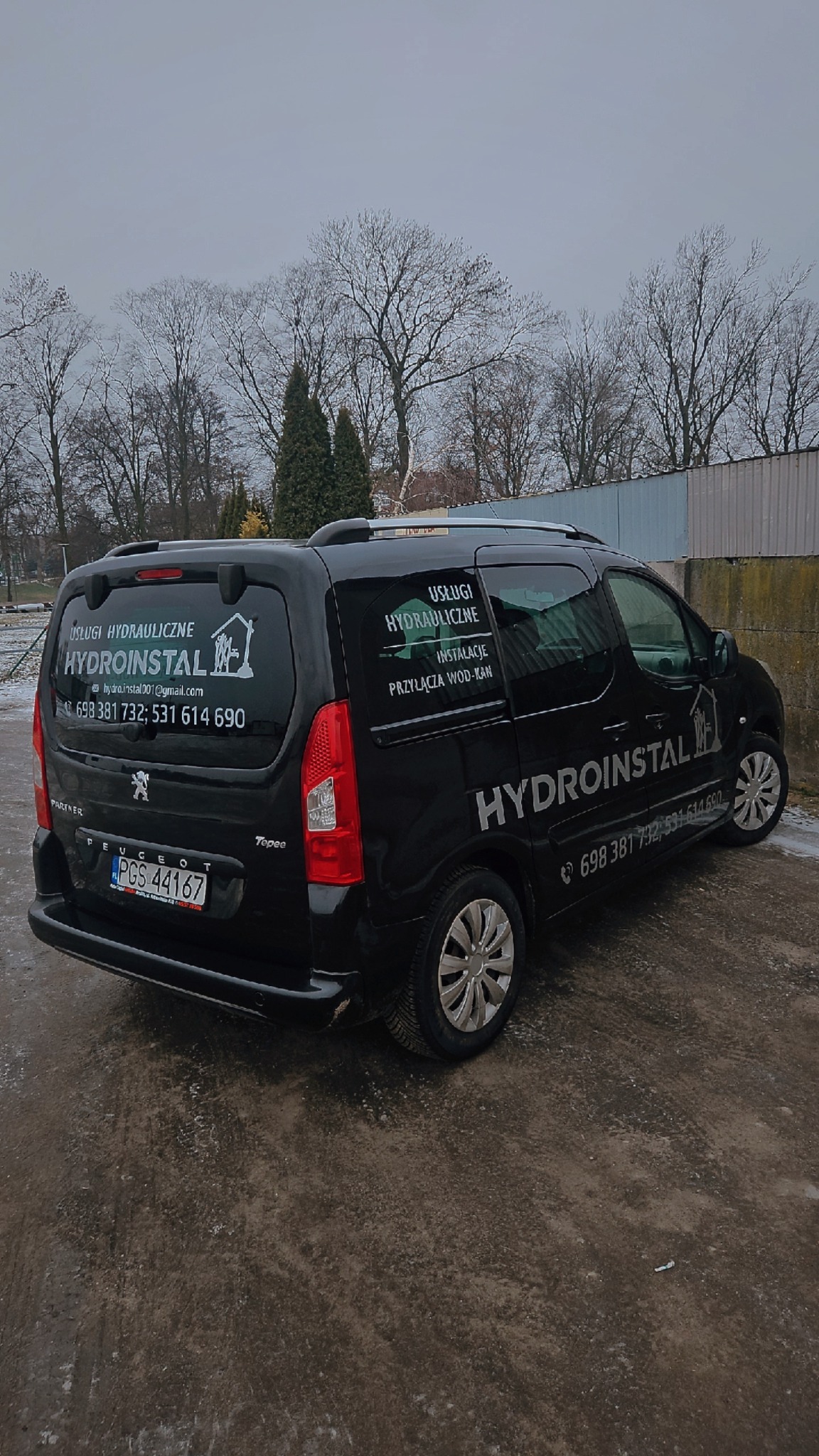 Czarny samochód dostawczy Peugeot Partner z logo i danymi kontaktowymi firmy hydraulicznej Hydroinstal, zaparkowany na zewnątrz w pochmurny dzień.